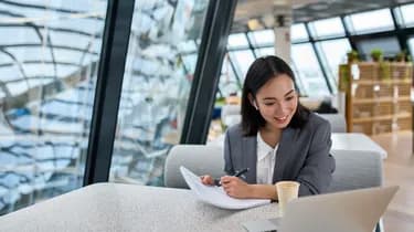 A woman in a gray blazer smiles while working on a laptop in a modern office, holding a pen and a notebook, with glass walls in the background.