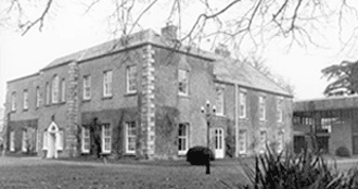 A large, historic brick building with white-trimmed windows and a gabled roof, surrounded by bare trees and a grassy area.