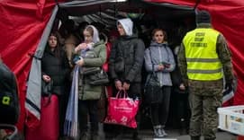 A group of Ukrainian refugees, bundled in warm clothing, stand under a red tent