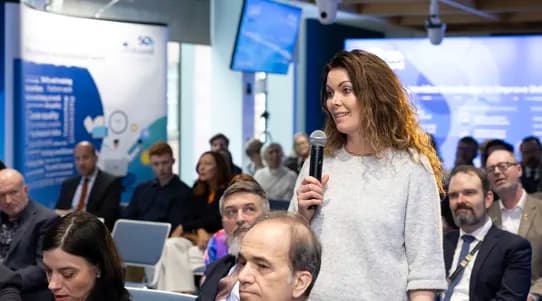 Woman in a conference setting speaking with microphone surrounded by other people