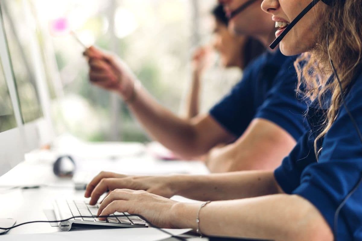 People working at computers in an office, wearing headsets and typing, with focus on a woman in the foreground.