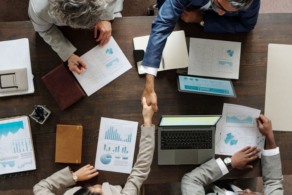 Top view showing group of middle aged multiethnic business professionals collaborating at table, two people shaking hands over laptop and financial charts, others reviewing documents